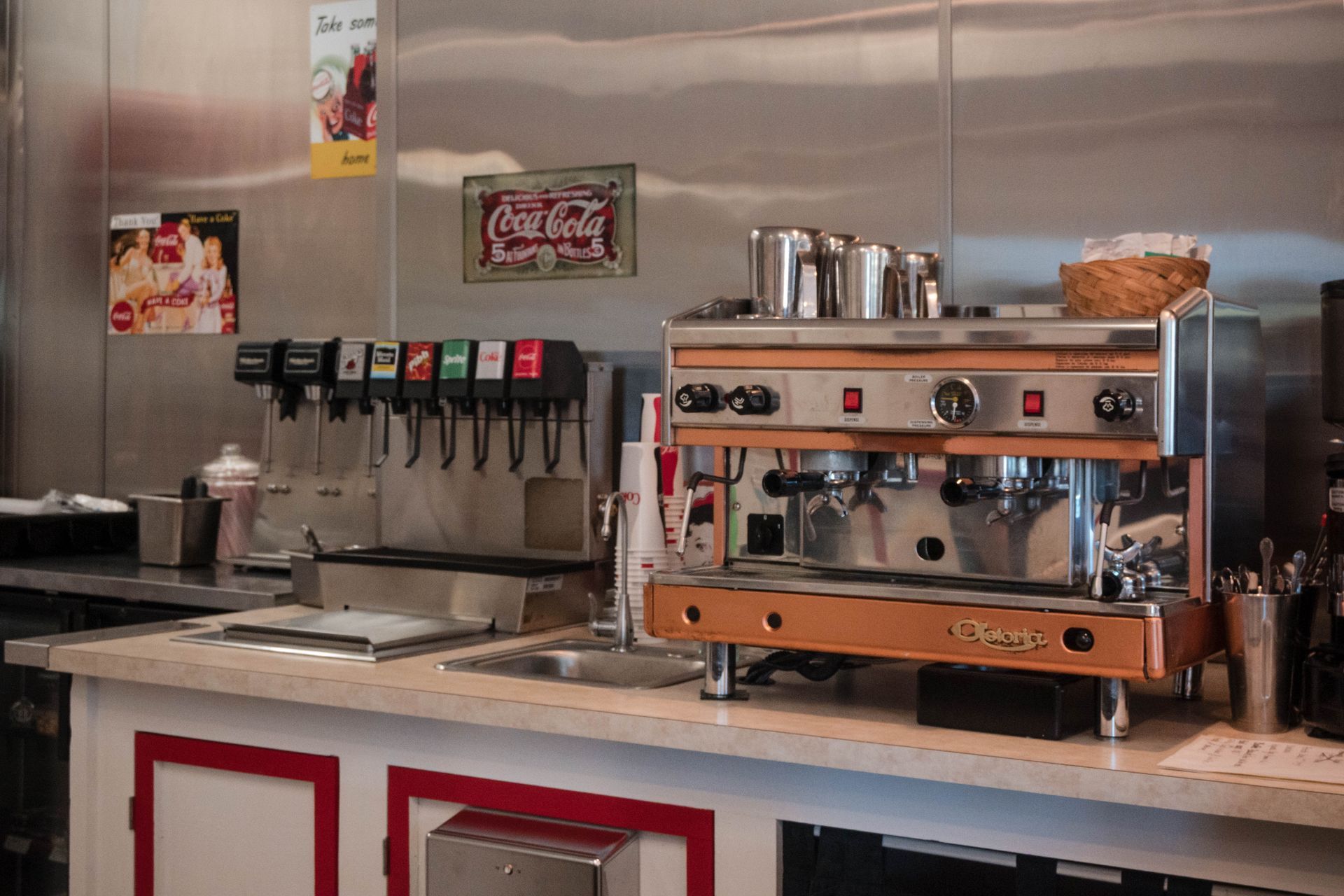 Coffee shop counter with espresso machine, drink taps, and stacked cups on a stainless steel bar.