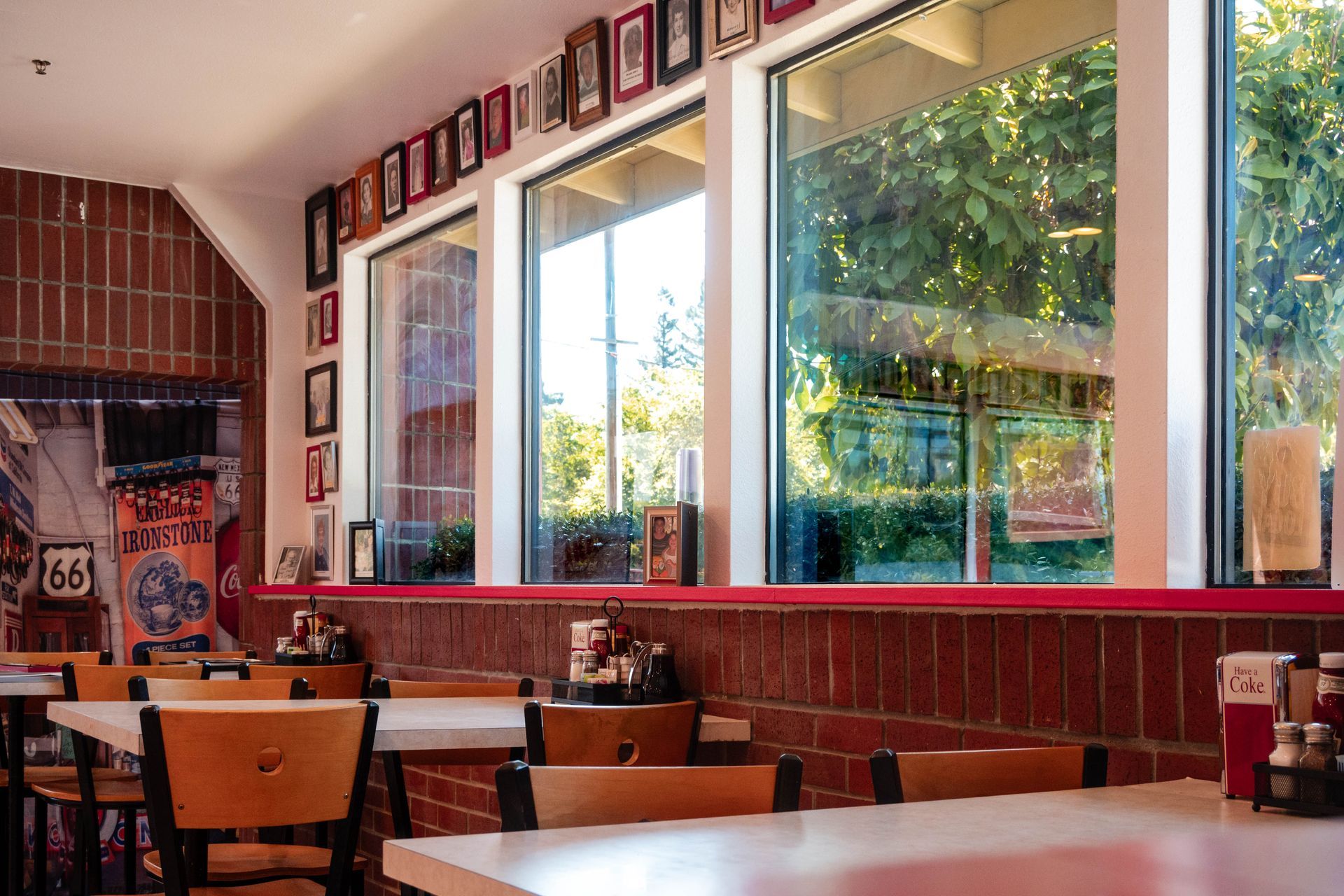 Empty restaurant dining area with red brick walls, wooden tables, and large sunlit windows overlooking greenery