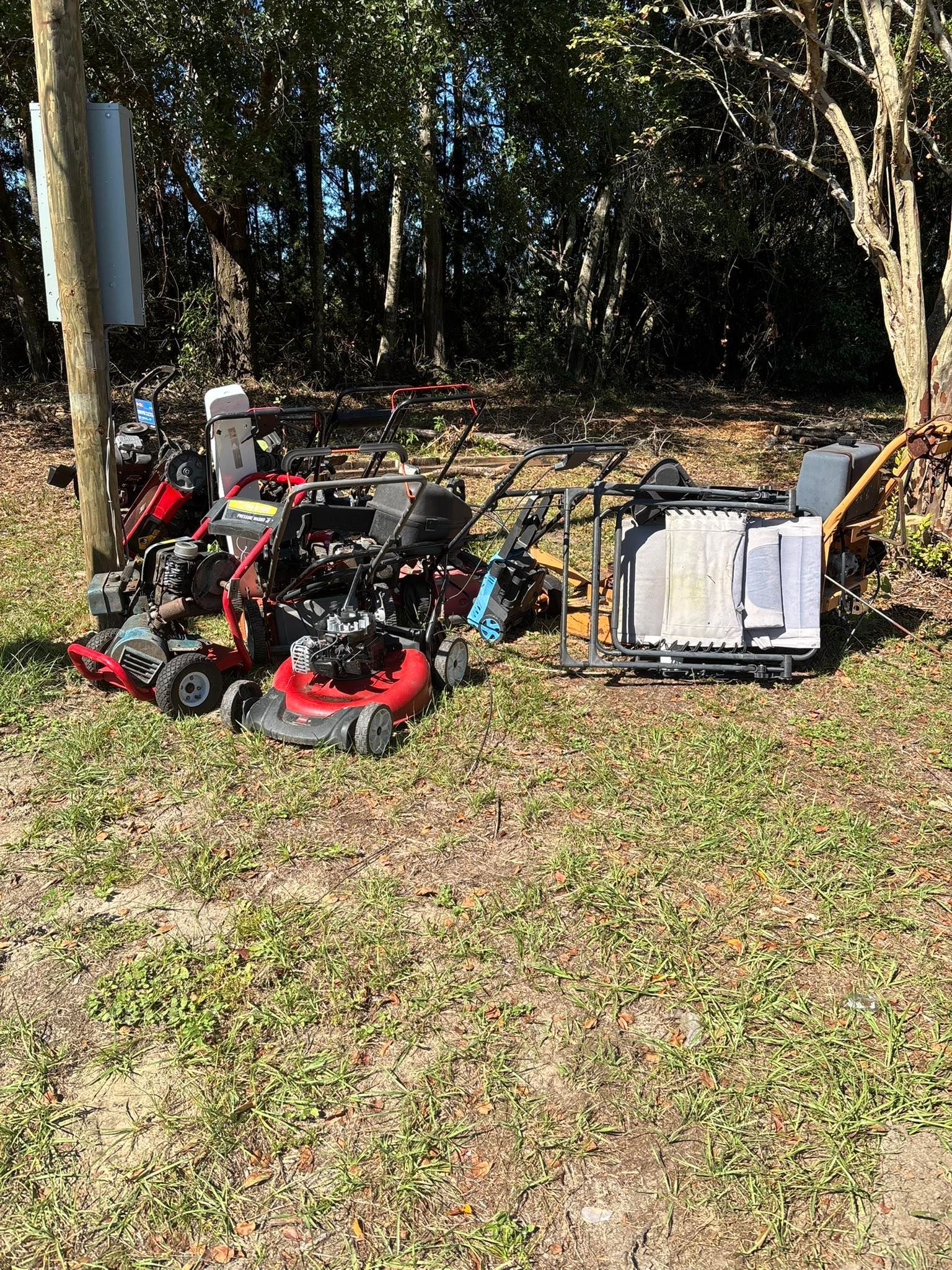 Pile of old lawn mowers and debris on a grassy area near trees.