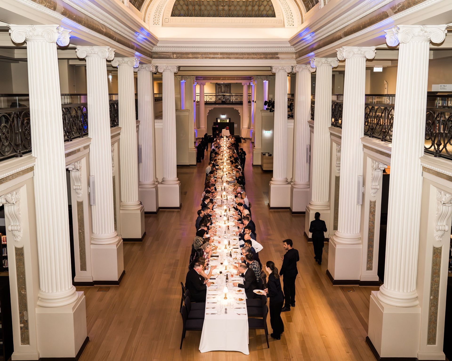 Elegant banquet hall with a long white-draped dinner table under tall white columns and a ornate ceiling.