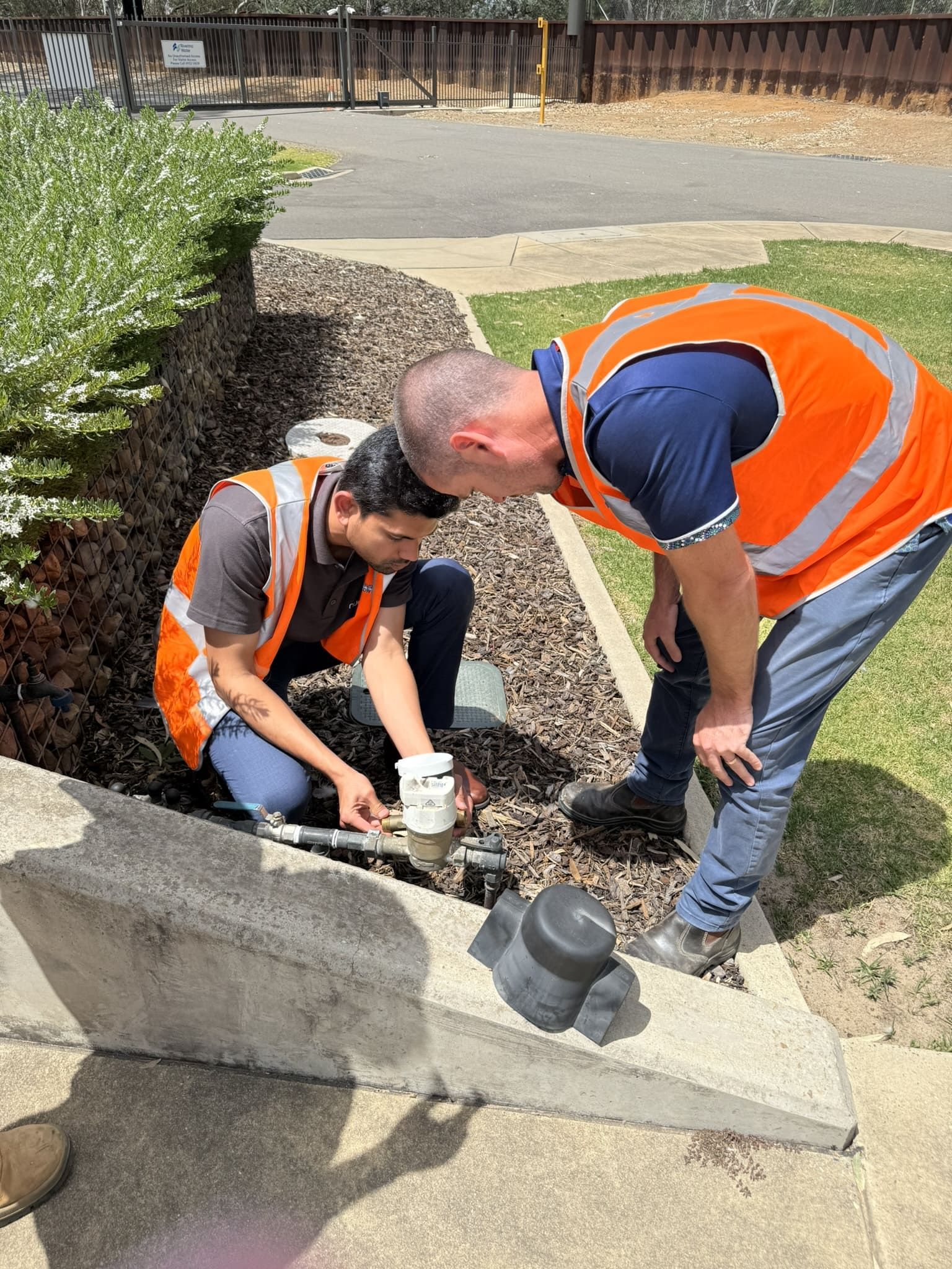 Two workers in orange safety vests inspecting a roadside drainage ditch outdoors.