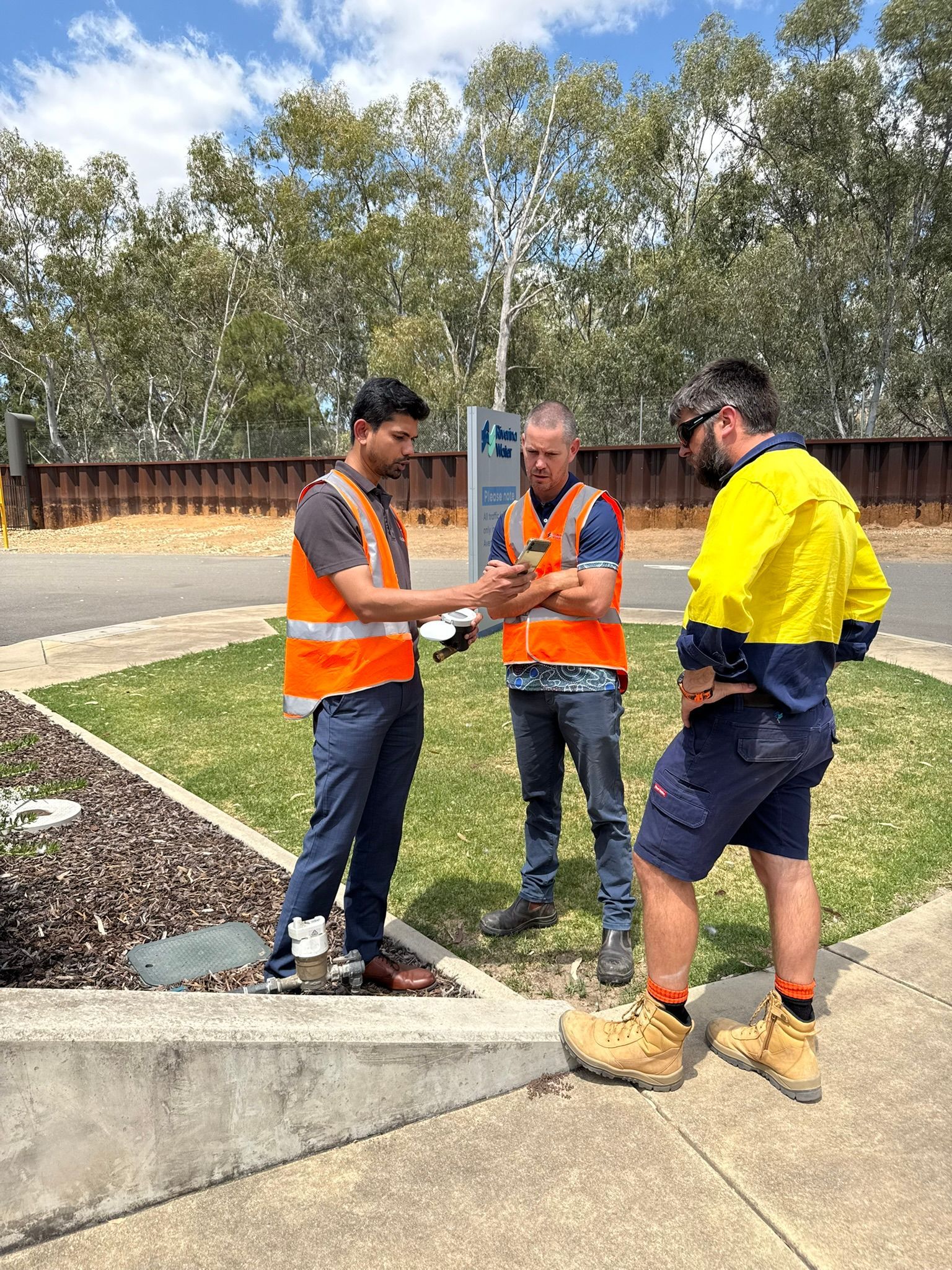 Three workers in safety gear discussing plans outdoors beside a curb and landscaped area.