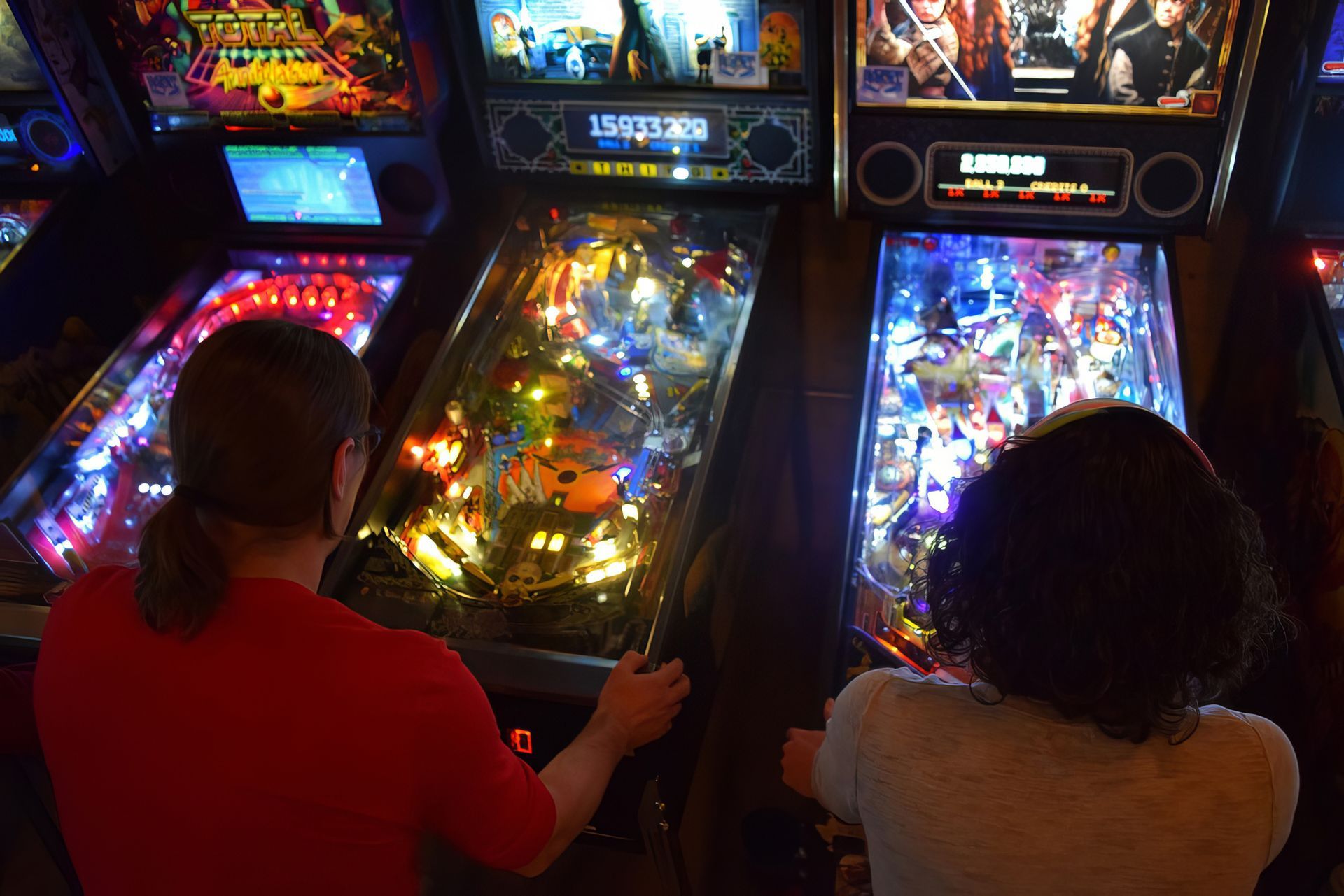 Two people are playing pinball machines in an arcade.