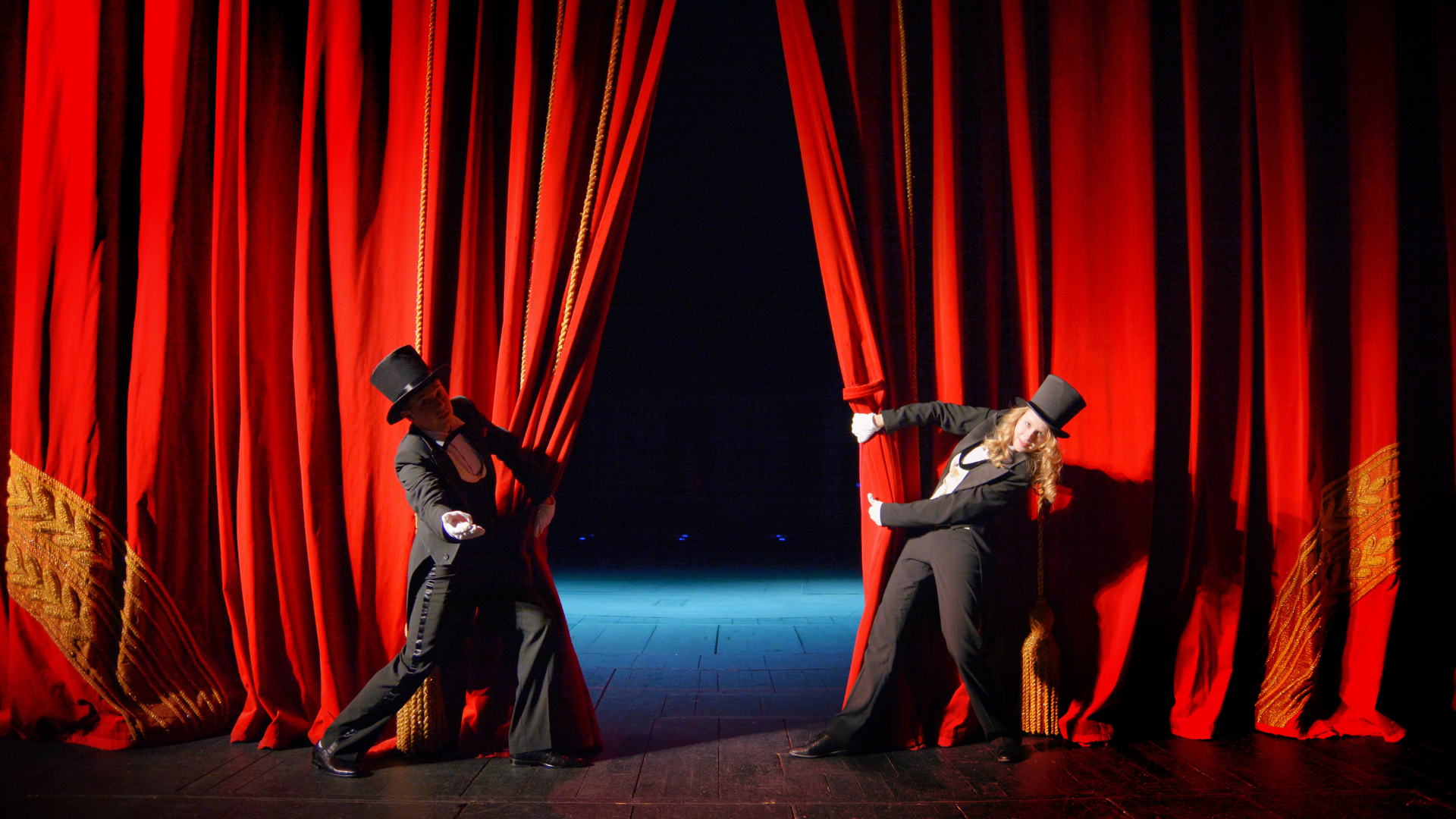 Two men in top hats are opening a red curtain on a stage.