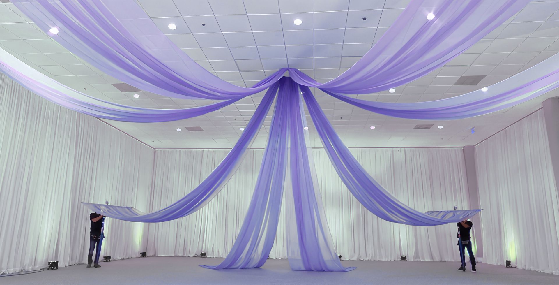 Two men are hanging purple fabric from the ceiling of a large room.