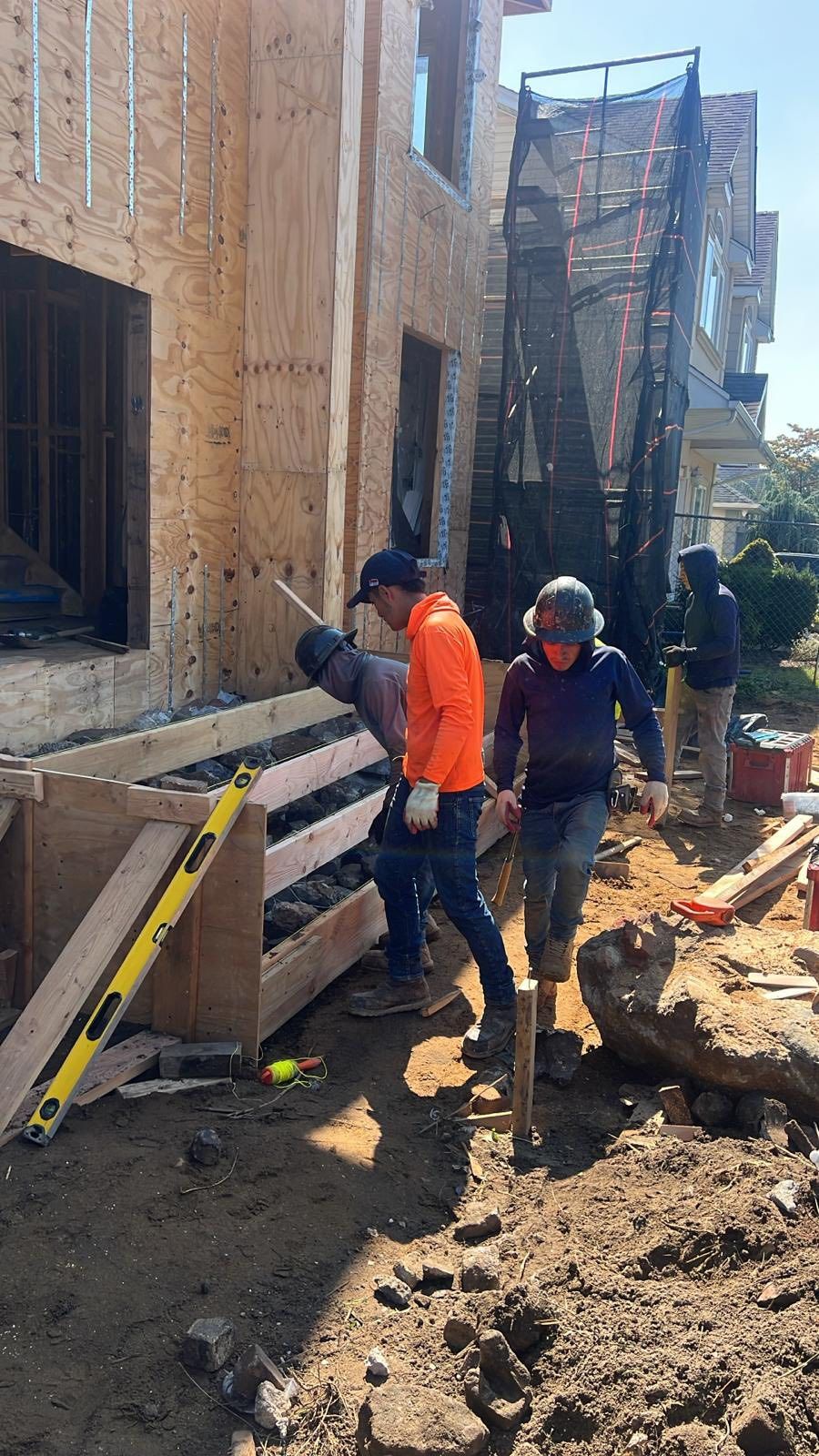 Construction workers building a foundation. Orange safety vests, lumber, dirt, and level are visible.
