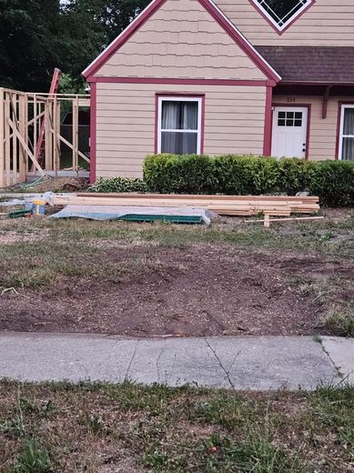 House under construction; wood framing, materials on the lawn. Beige exterior, red trim, bushes, sidewalk.