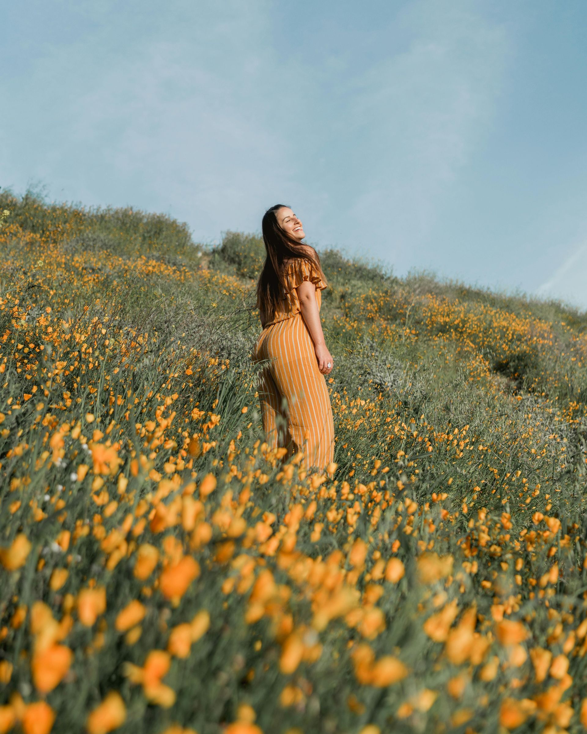 Woman in yellow dress smiles on a hillside covered in orange wildflowers, blue sky.