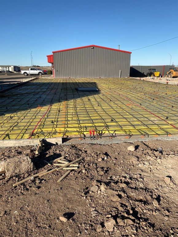 A construction site shows a yellow vapor barrier and metal rebar grid prepared for a concrete pour in front of a shed.