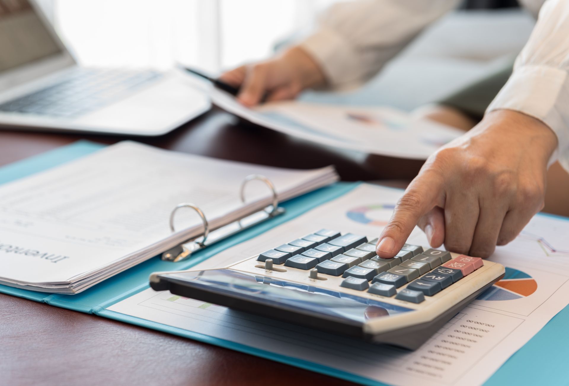 Person using calculator with documents, laptop, and graphs on a desk.