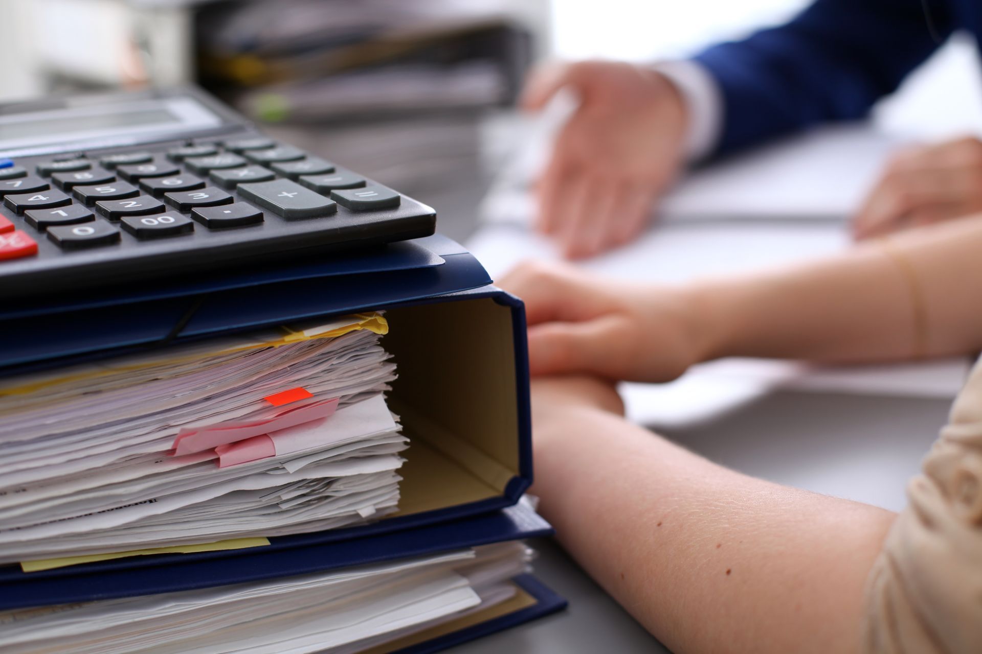 Pile of papers with calculator; people discussing documents in office.