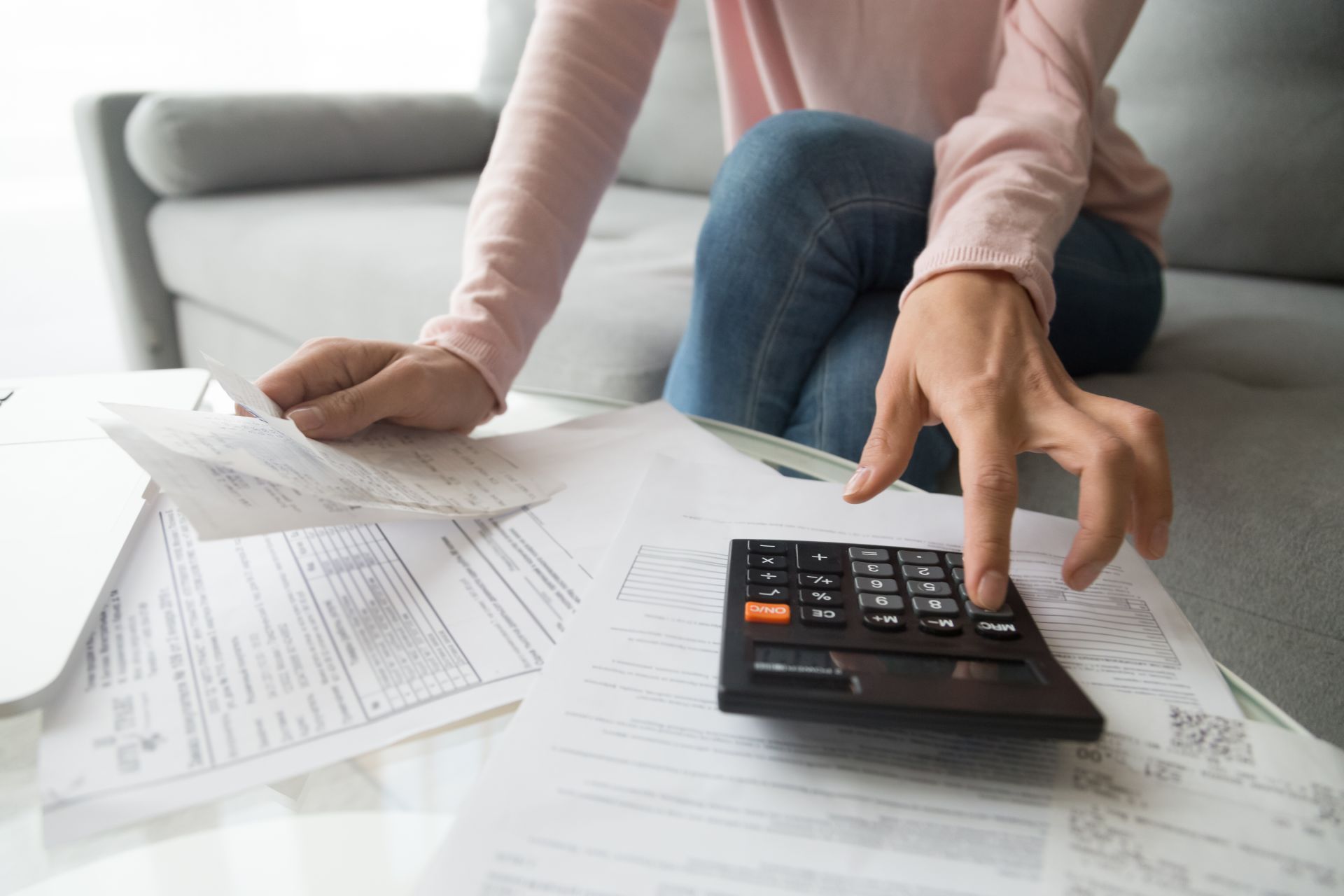Woman calculating bills on a calculator while sitting on a couch, surrounded by papers.