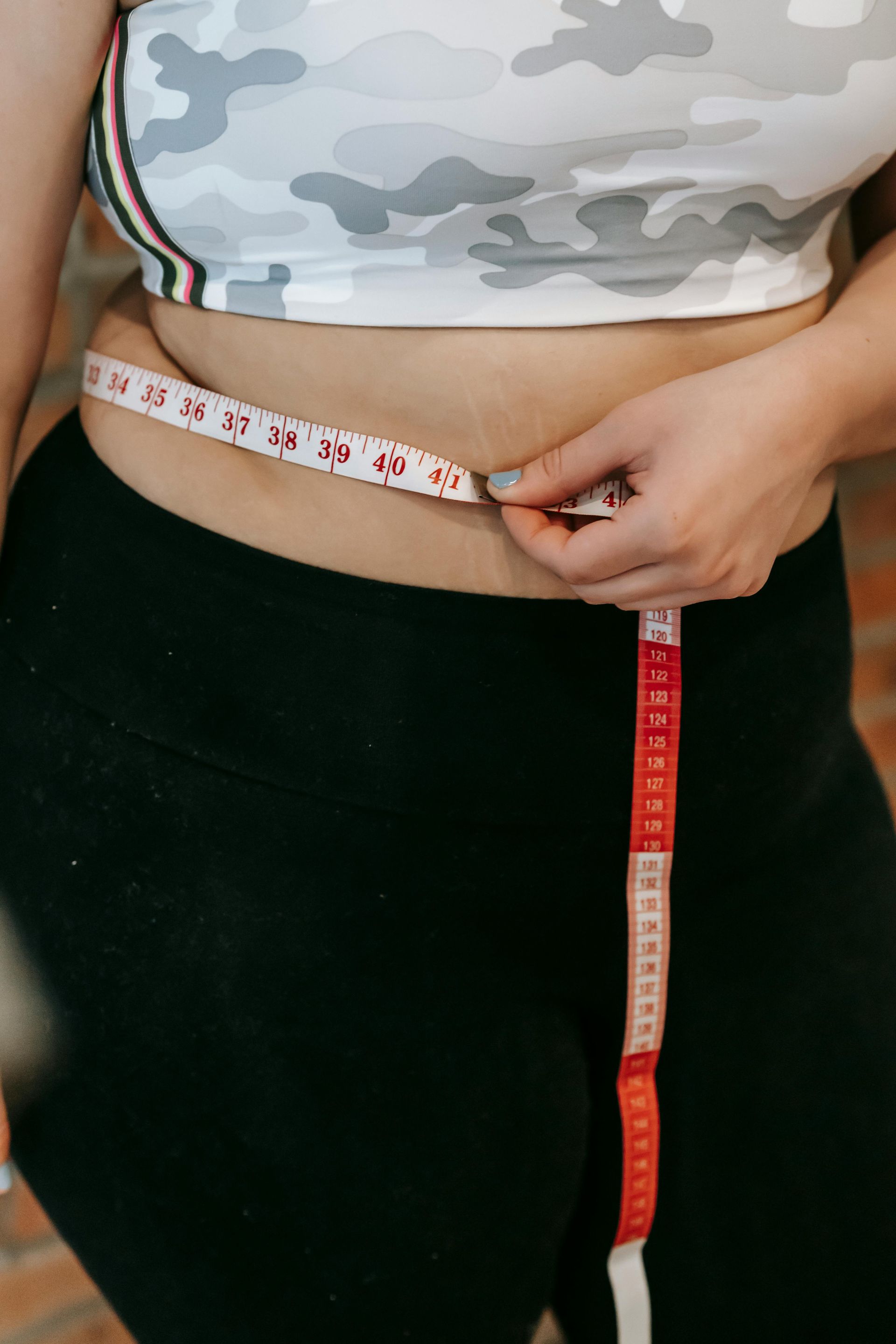 Person measuring their waist with a red and white measuring tape. They wear a camo crop top and black leggings.