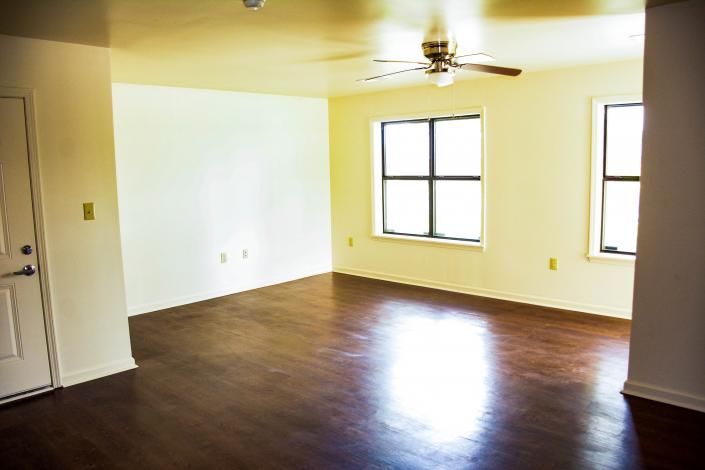 Empty living room with dark wood floor, white walls, ceiling fan, and two windows.