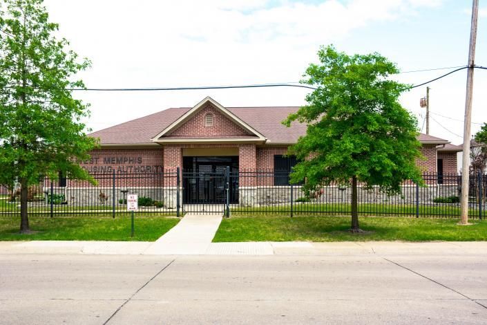 Brick house with a fenced front yard, walkway, and trees along a street