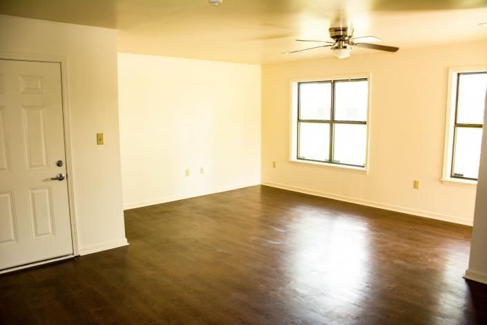Empty living room with dark wood floors, white walls, ceiling fan, and two windows.