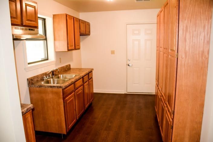 Small empty kitchen with wood cabinets, stainless sink, dark floor, and white door.