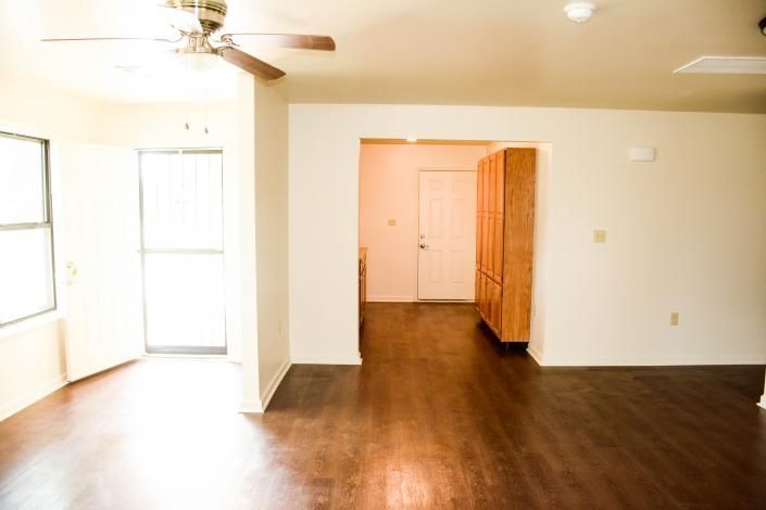 Empty living room with dark wood floors, ceiling fan, and bright windows leading to a hallway