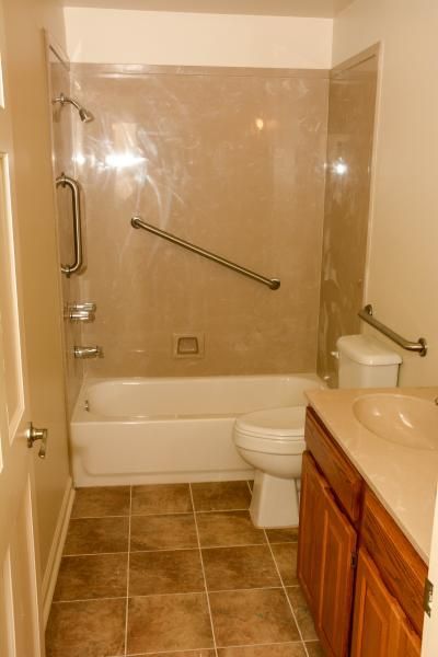 Accessible bathroom with tub, toilet, tan tile walls, and grab bars