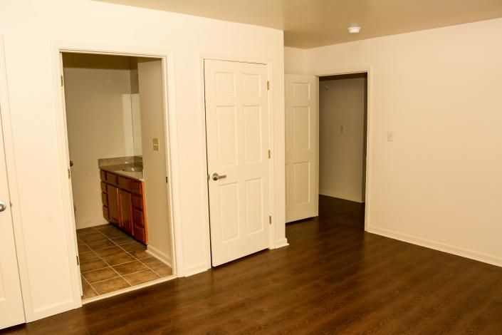 Empty room with dark wood floor, white walls, and open doorways to a kitchen and hallway