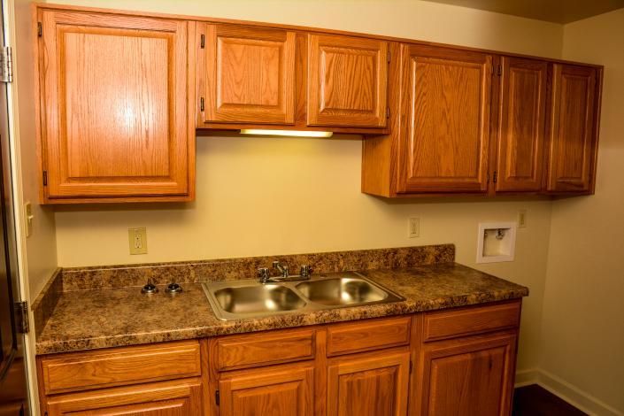 Wooden kitchen cabinets and granite countertop with double sink and beige backsplash under warm lighting