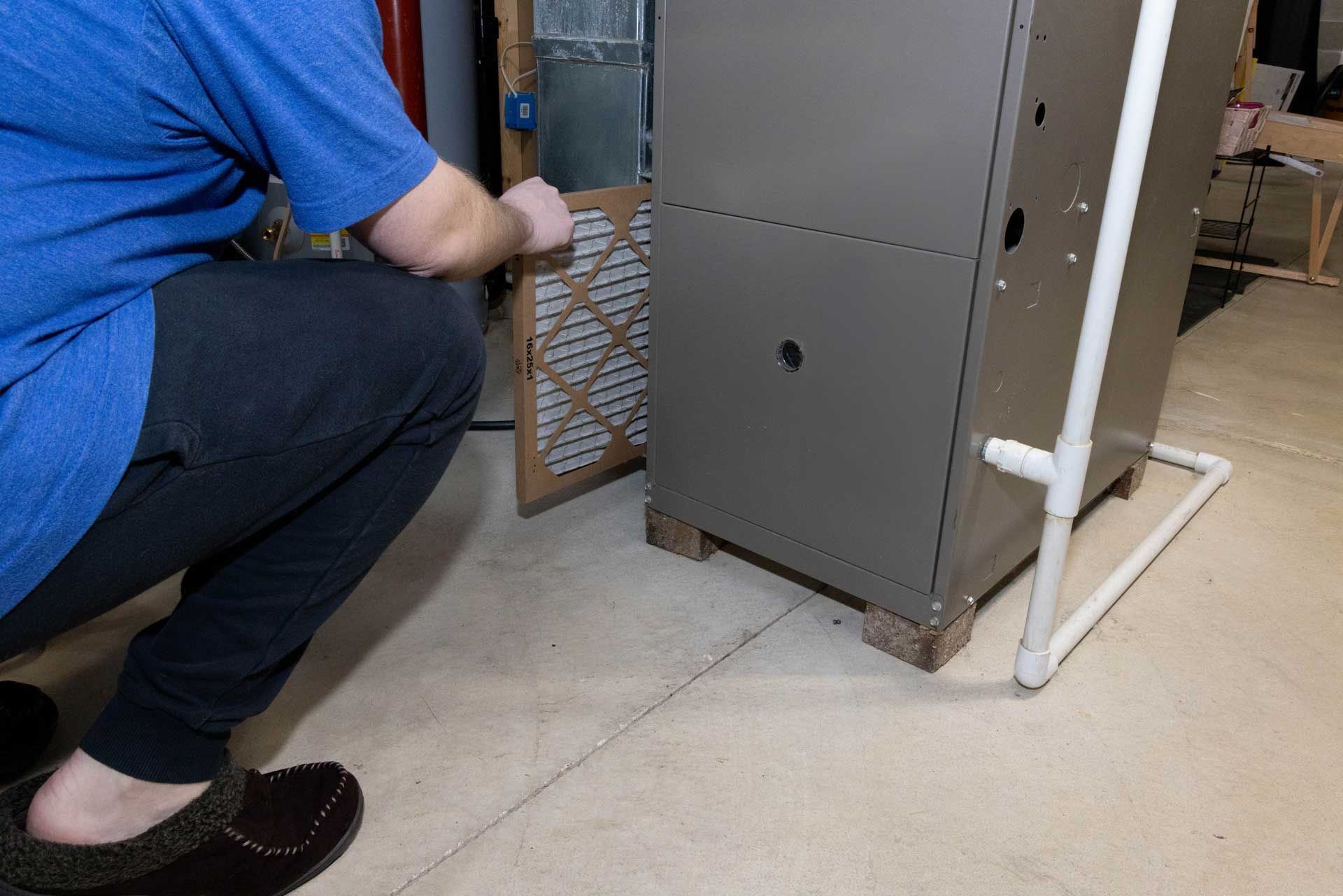 A person in a blue shirt holds a rectangular air filter while removing or inserting it into a home furnace unit.