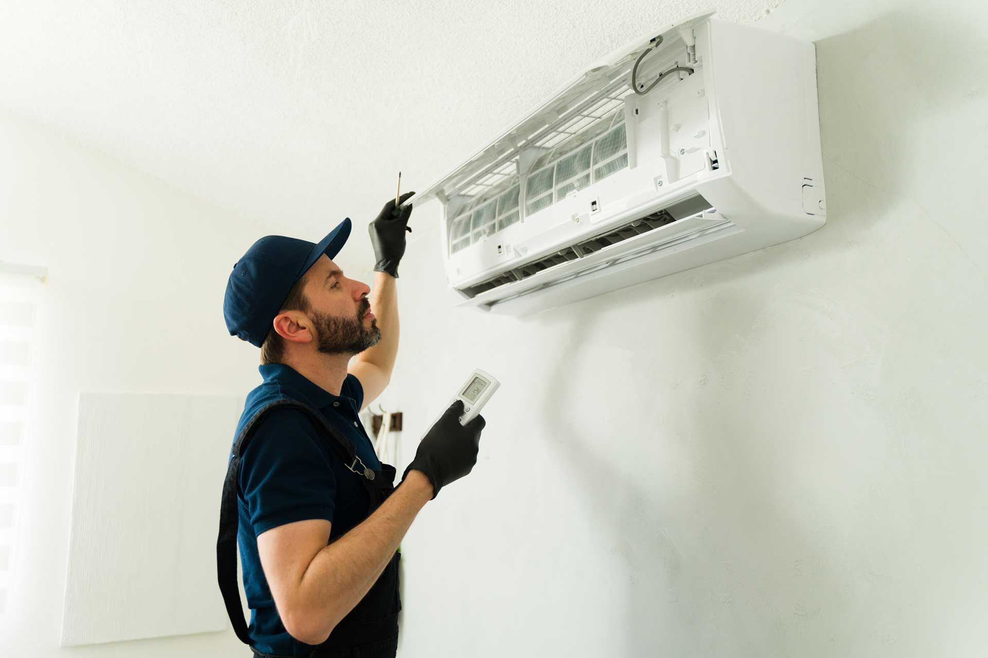 A technician in a blue uniform and gloves inspects a wall-mounted air conditioner while holding a remote control.