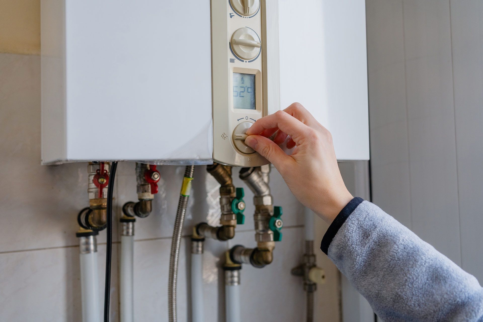A person's hand adjusts a dial on a white wall-mounted boiler unit with visible pipes underneath.