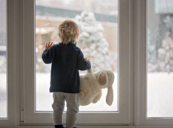 A little boy is holding a stuffed animal and looking out of a window.