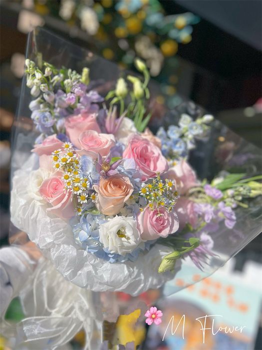 一束粉色、白色和紫色的花朵，用透明塑料包裹，背景是模糊的户外景色。