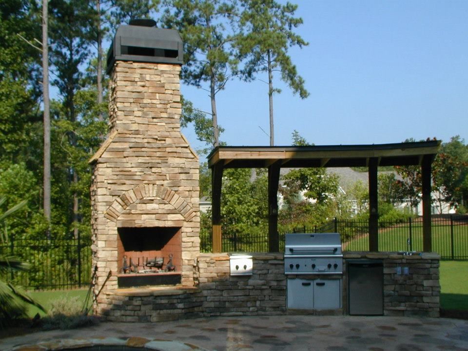 Outdoor stone fireplace and grilling station with a roof, in a backyard setting.