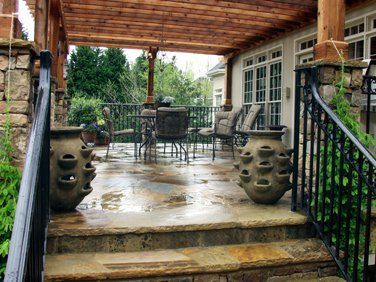 Stone patio with pergola, potted plants, and wrought iron railing. Patio furniture visible in the background.