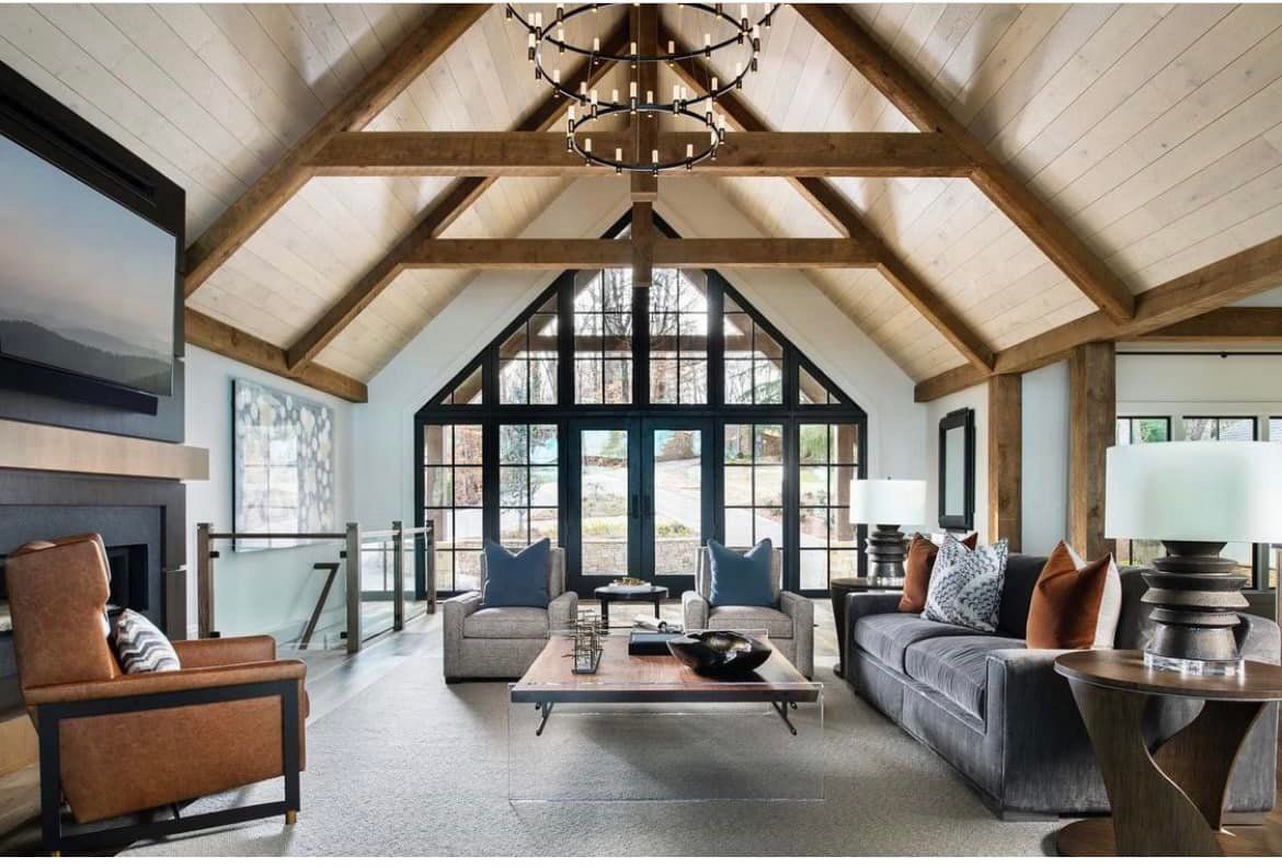 Living room with vaulted ceiling, exposed beams, large windows. Gray sofa, chairs, fireplace.
