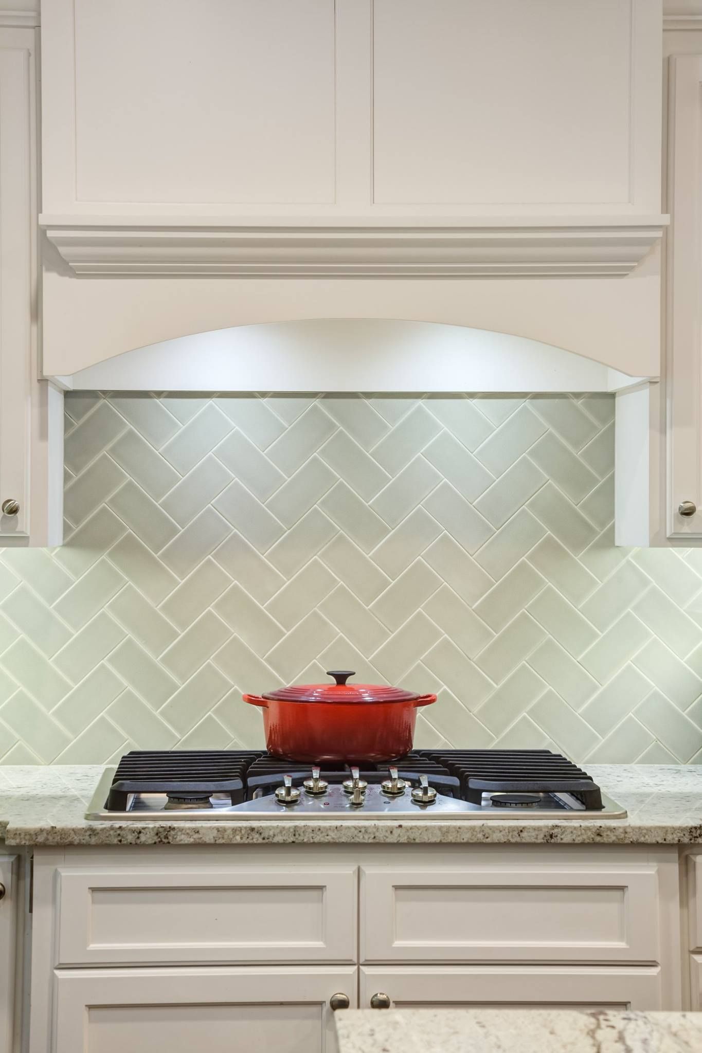 Red pot on a stovetop in a kitchen with white cabinets and herringbone tile backsplash.