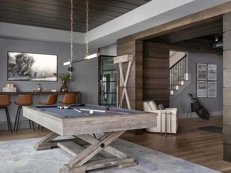 Pool table in a finished basement with bar, seating, and golf club display. Gray and wood tones.