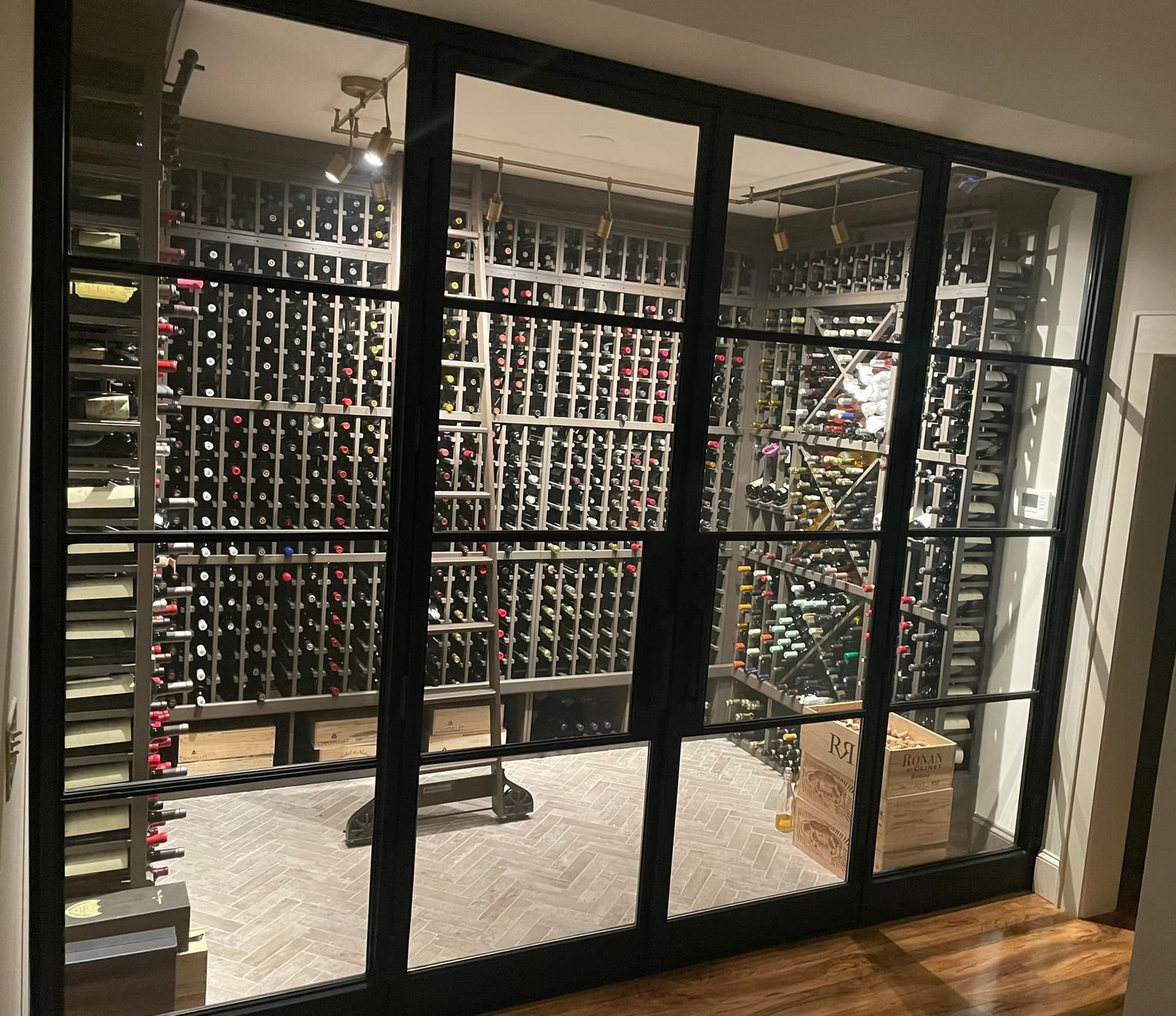 Wine cellar with glass doors, filled with bottles on racks; a metal ladder is inside.