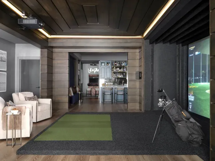Golf simulator room with green turf, golf bag, and screen displaying golf course. View through a doorway to a bar.