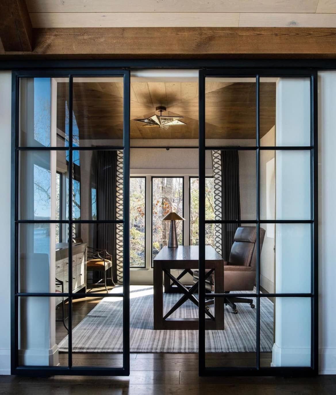 Sliding glass doors reveal a home office with a desk, chair, and windows; wood beams overhead.