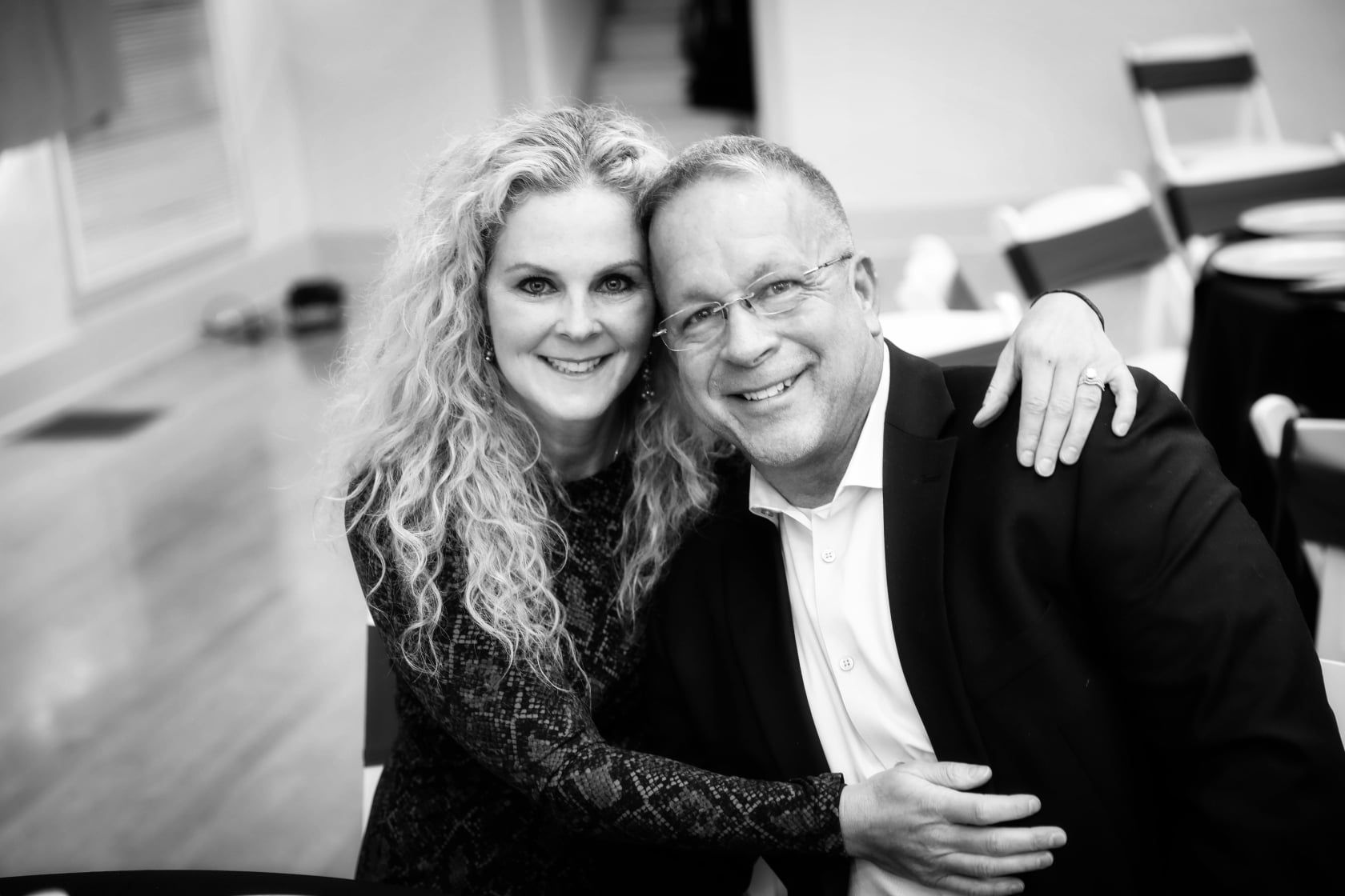 Smiling couple embraces, woman's arm around man's shoulders. Black and white photo, indoors, event setting.