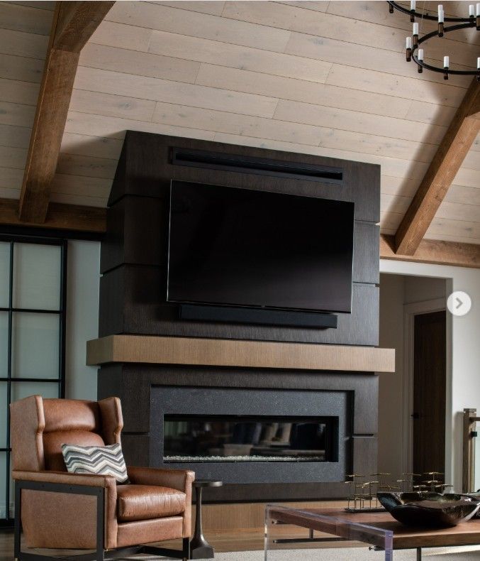Modern living room with fireplace, TV, and leather armchair. Dark wood mantel, light ceiling, and large windows.