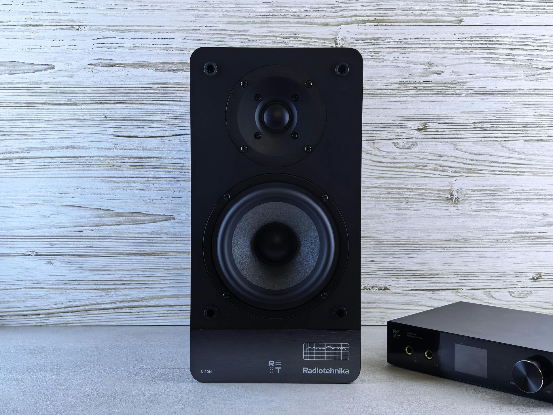 Black speaker and audio amplifier on a gray surface against a white wood-paneled wall.