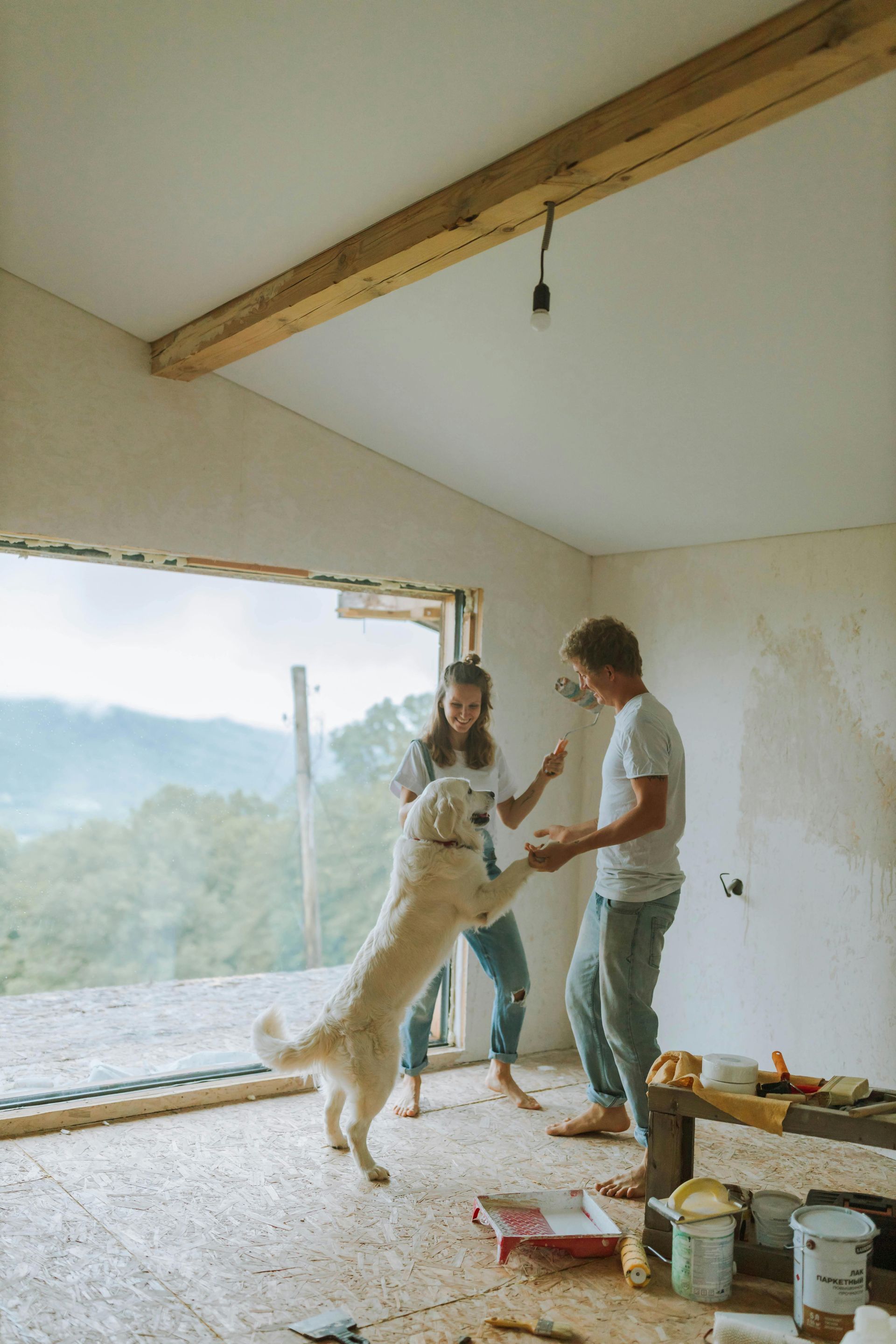 Couple and golden retriever playing in unfinished room with window overlooking trees.