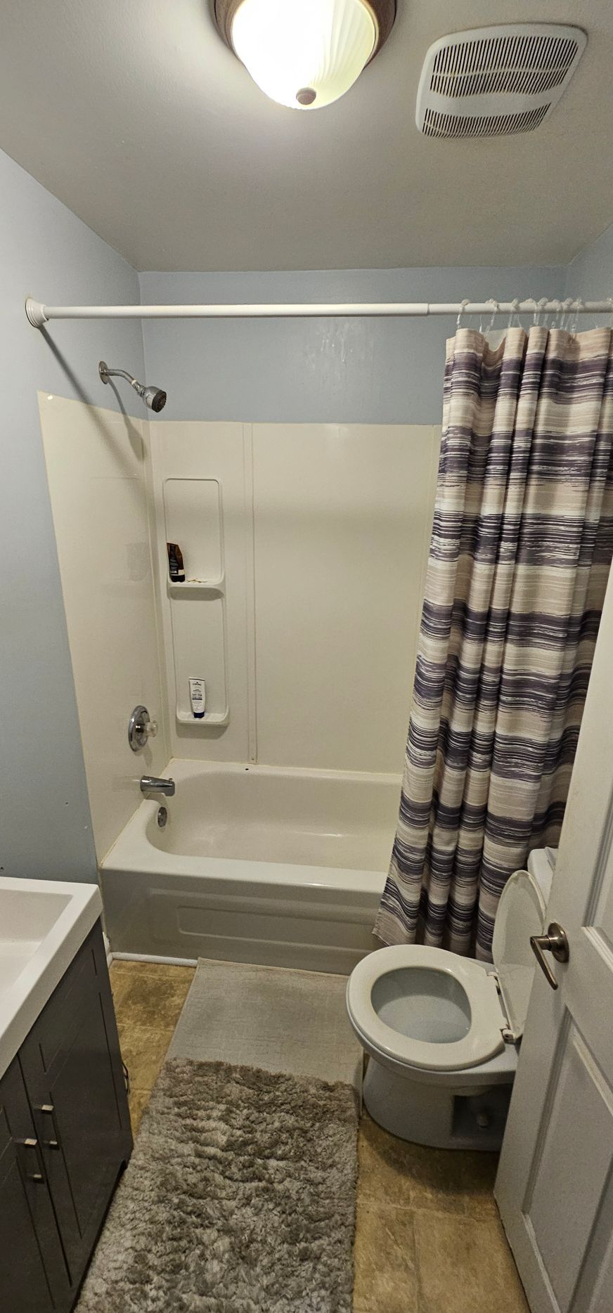 A small bathroom with a bathtub, toilet, and vanity. There's a striped shower curtain and a shaggy rug.