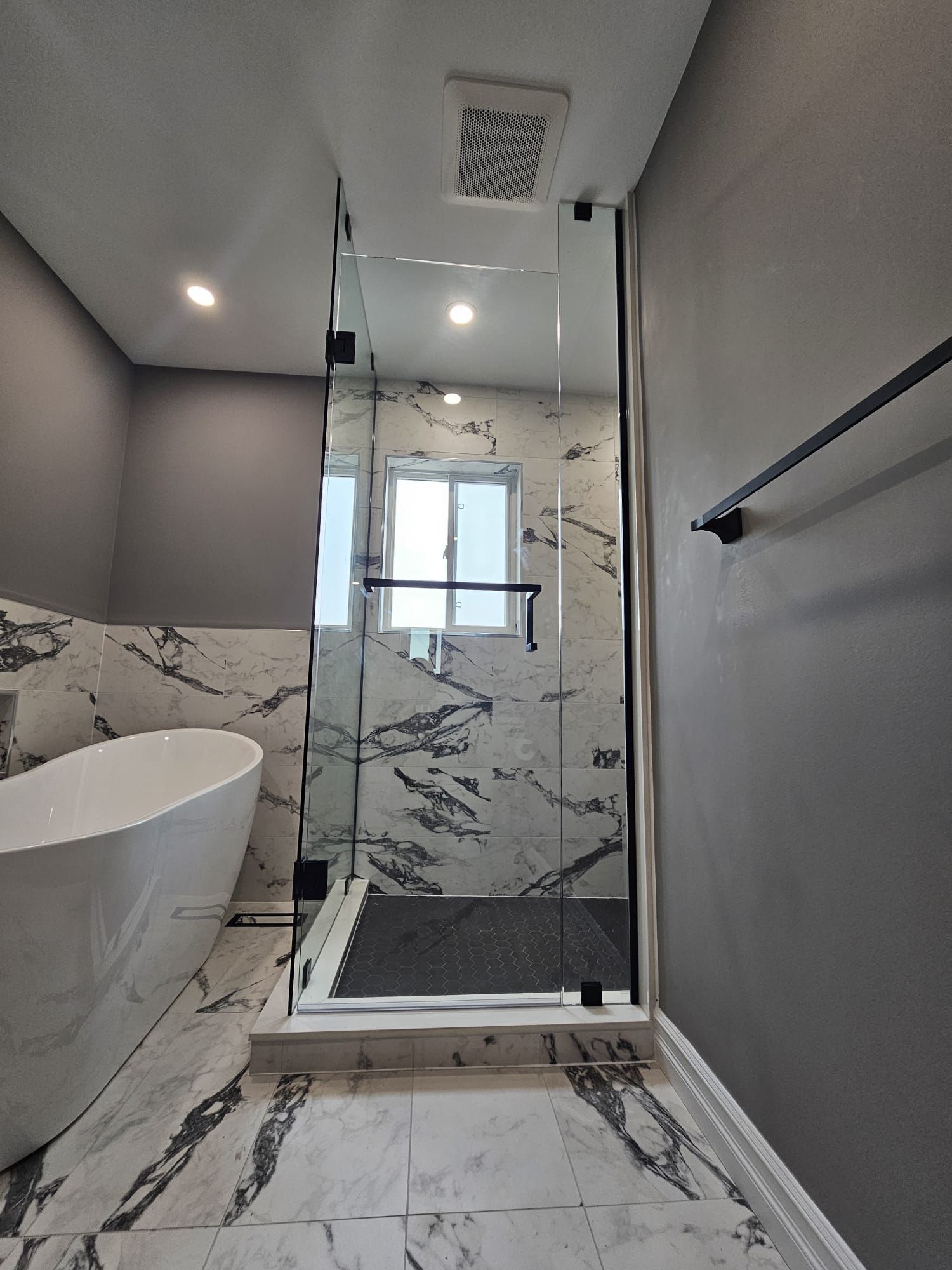 Modern bathroom with a glass shower, marble tile, and freestanding tub. Gray walls and dark fixtures.