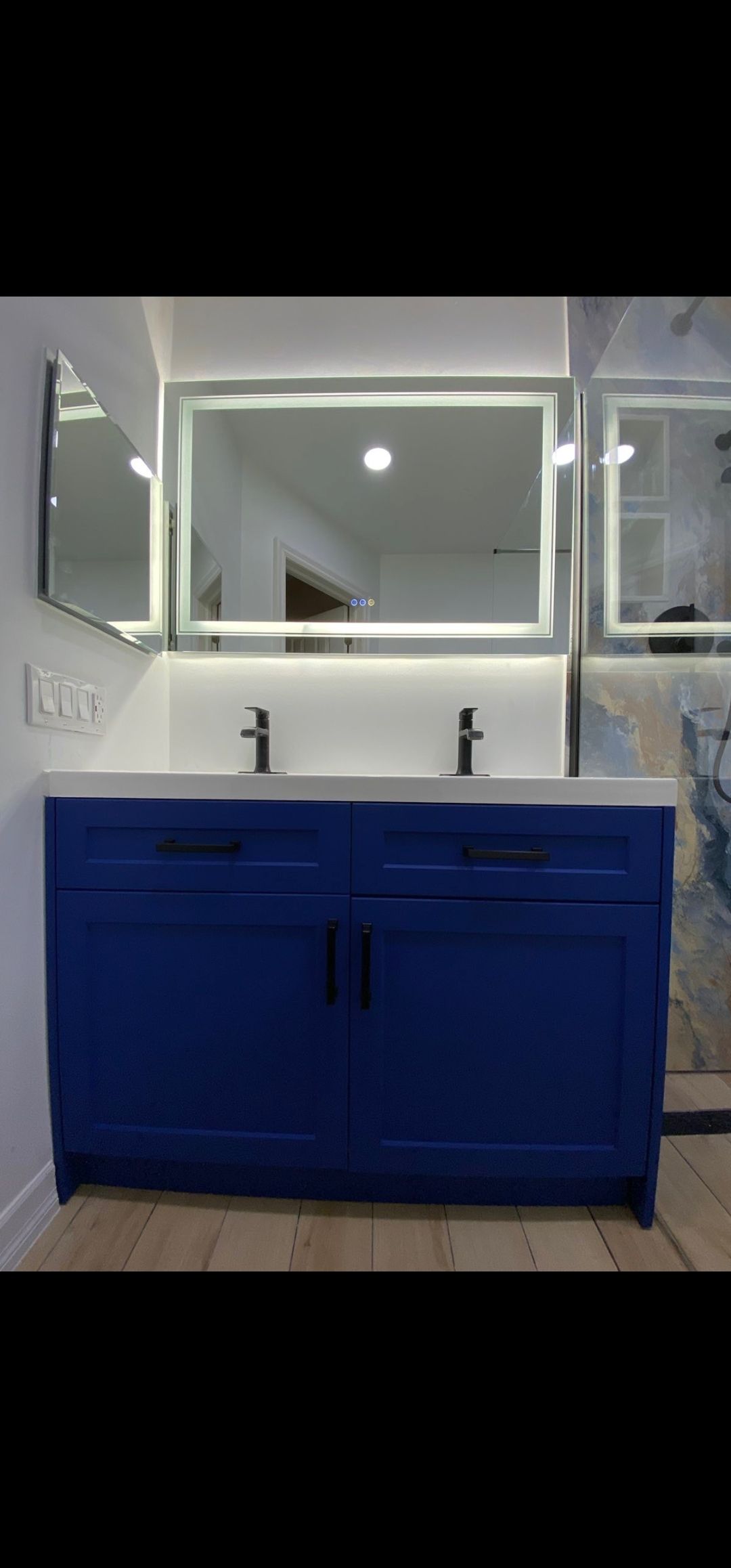 A blue bathroom vanity with a white countertop and two faucets, under a lit-up mirror.