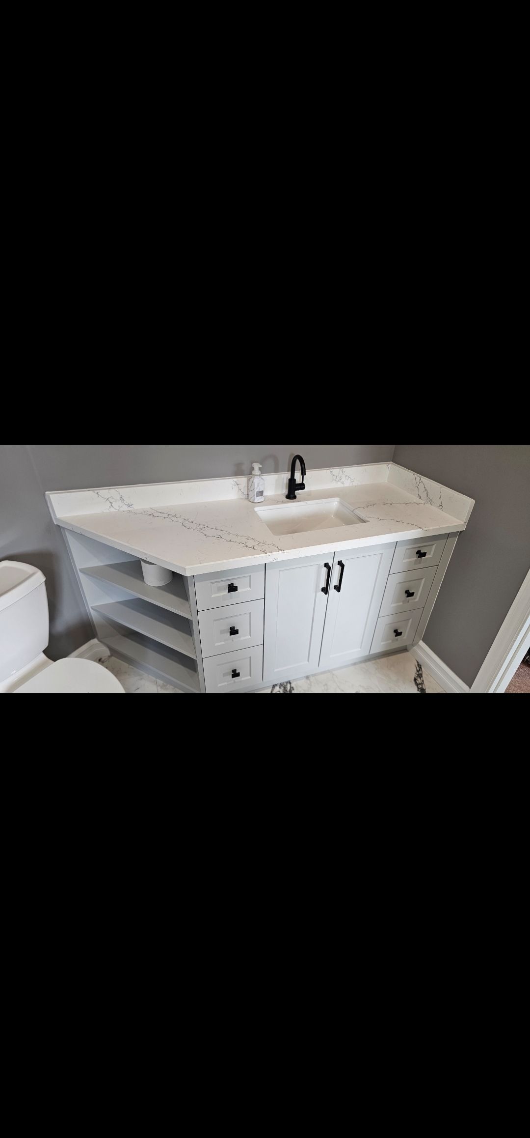 Bathroom vanity with a white countertop and black faucet. It has open shelves and a cabinet.