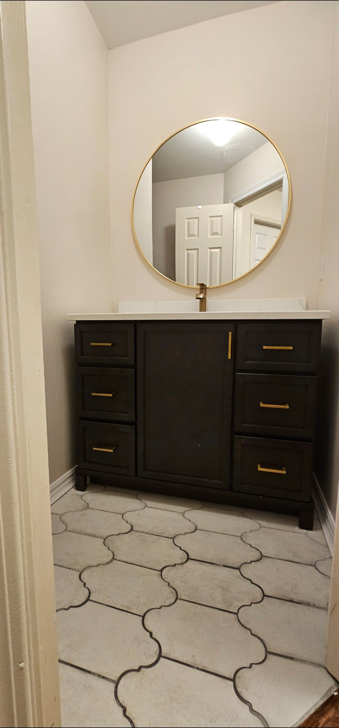 A black bathroom vanity with gold hardware and a round gold mirror, the floor is white tile with black trim.