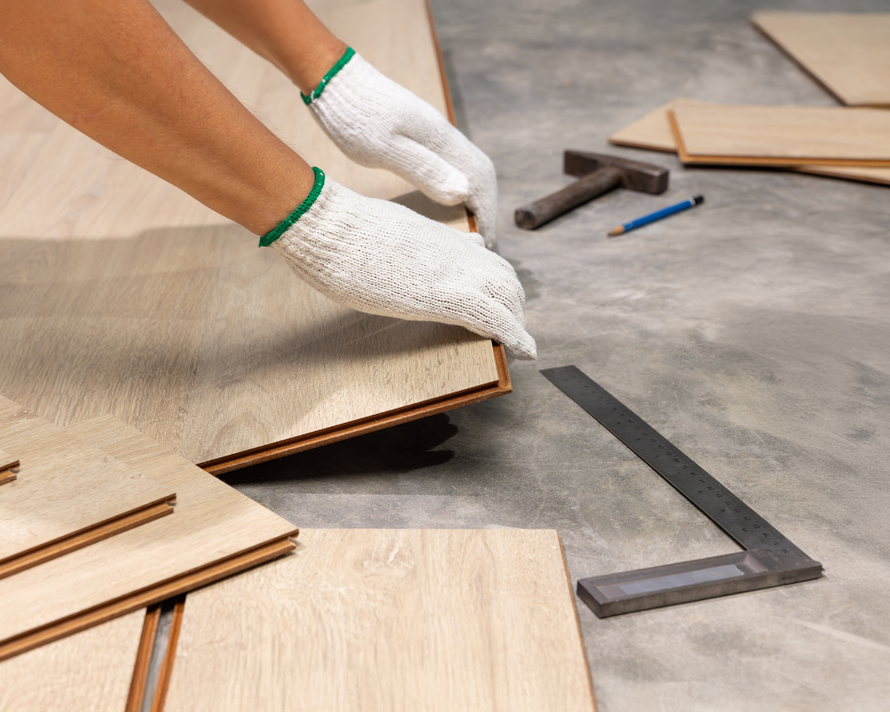 Hands in gloves installing laminate flooring, with tools on concrete floor.