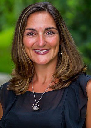 Woman with brown hair, smiling, wearing a black top and silver necklace, outdoors.