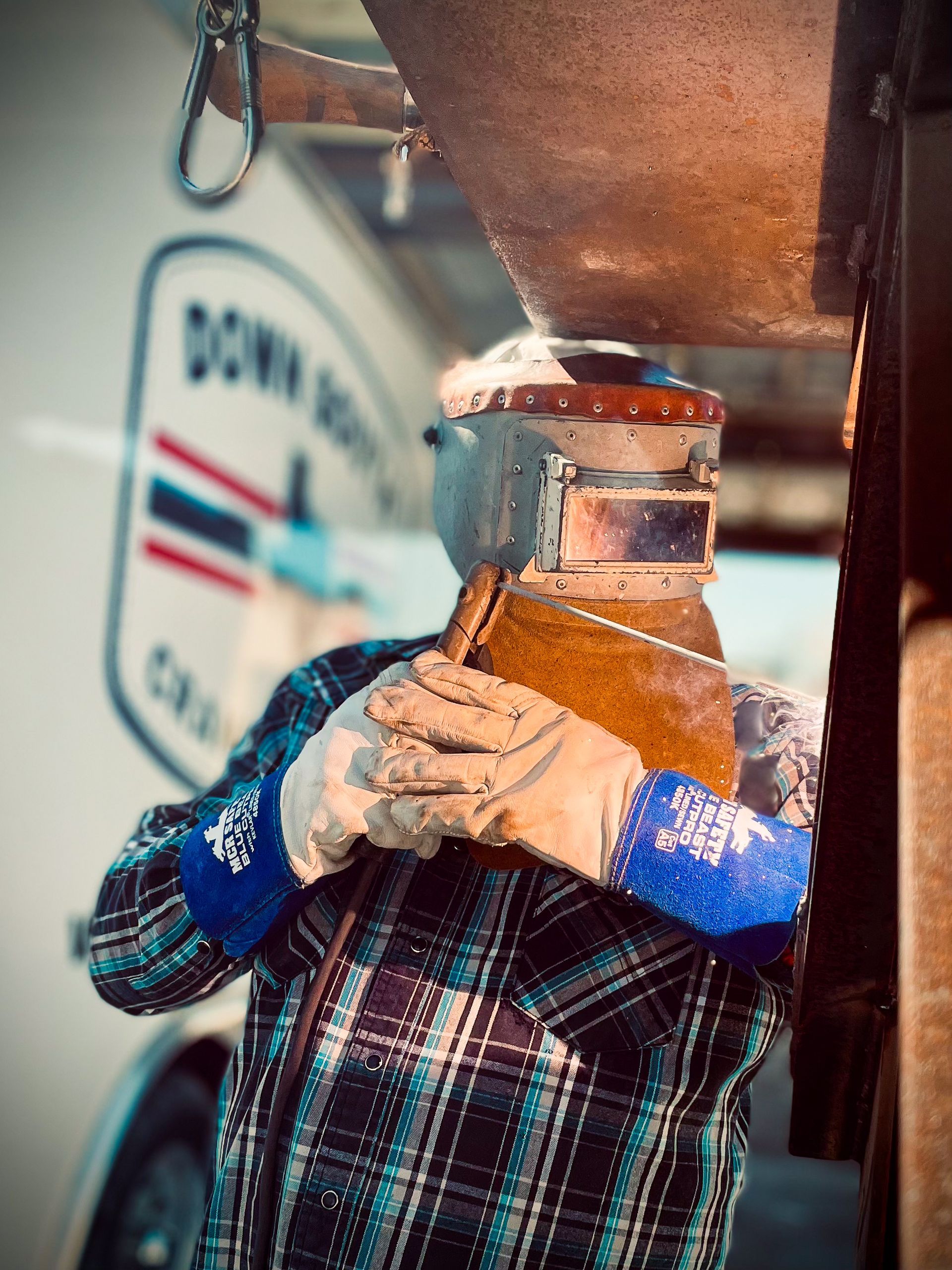 Welder in a plaid shirt with a welding helmet and gloves, working on equipment near a white truck.
